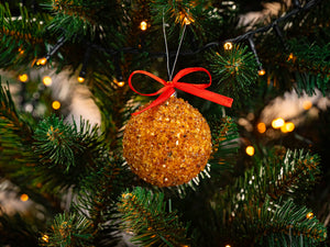 BalticBliss genuine Baltic amber Christmas tree ornament ball crafted from sparkling natural amber chips and topped with a bright red ribbon bow, hanging on lush green fir branch among warm twinkle lights, handmade in Lithuania, eco‑friendly authentic amber holiday décor ships worldwide