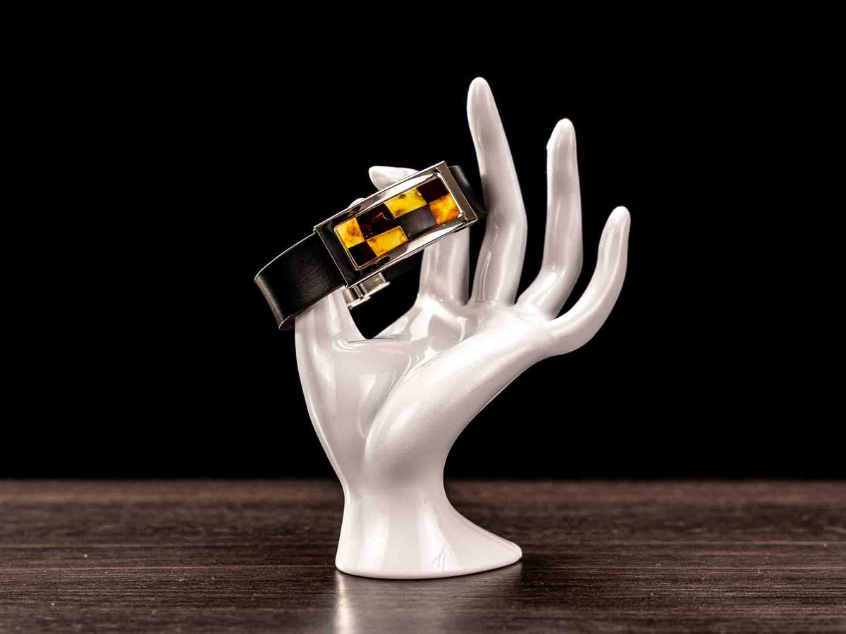 Wide black leather bracelet with a rectangular amber mosaic inlay, displayed on a white ceramic hand against a black background.