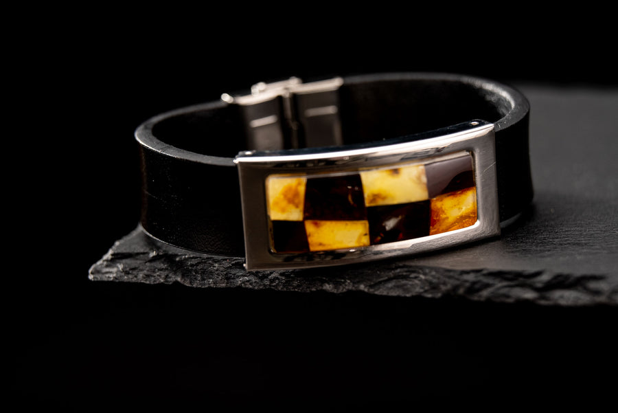 Wide black leather bracelet with a rectangular mosaic of light and dark amber pieces in a silver-coloured frame, displayed on dark slate.