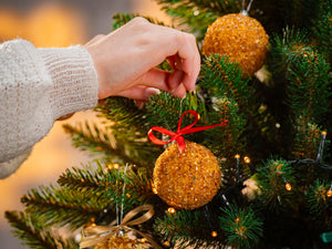 Hand placing BalticBliss genuine Baltic amber Christmas tree ornament ball with shimmering natural beads and red ribbon on lush green fir branch, handmade in Lithuania, authentic amber holiday décor ships worldwide