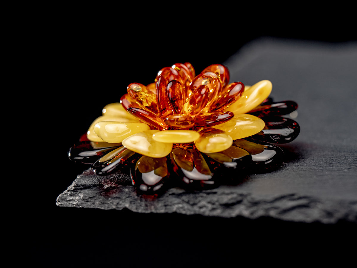Amber flower brooch and pendant viewed from a low side angle on a dark stone surface.