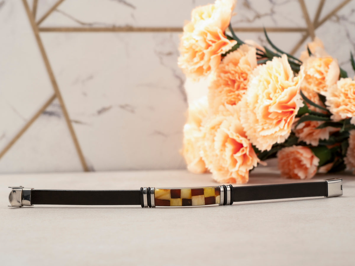 Handcrafted black genuine leather bracelet with real Baltic amber mosaic inlay laid flat on white surface with peach carnations and marble background – luxury unisex jewelry lifestyle photo.