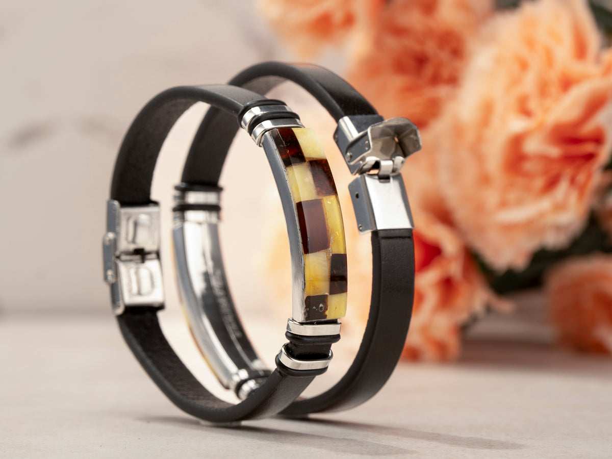 Two handcrafted black genuine leather bracelets with real Baltic amber mosaic inlays stacked together in a luxury lifestyle shot. Unisex amber jewelry with silver-tone hardware against soft peach floral background – perfect for wearing as a matching pair or set.