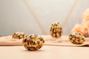 Premium studio lifestyle photo of four handcrafted genuine amber mosaic Easter eggs on soft peach fabric. One egg in foreground, others on clear stands with gentle peach flowers in background – luxury minimalist Easter home decor.